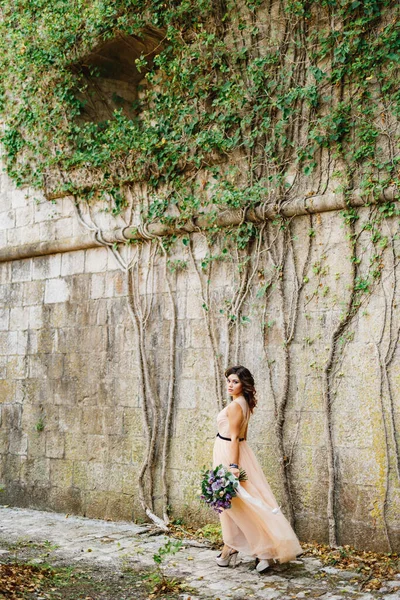 Bella sposa in un abito pastello passeggiate con un lussuoso mazzo di fiori lungo il muro di pietra — Foto Stock