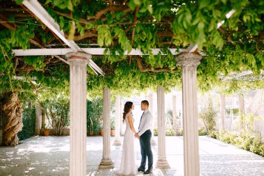Mutlu gelin ve damat, güneşli bir günde güzel yeşil bir bahçede sütunların arasında el ele tutuşuyorlar..