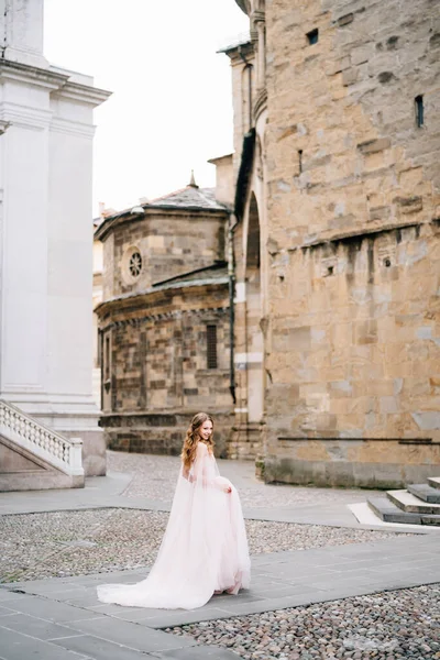 Bela noiva fica em um vestido longo sobre as pedras de paralelepípedos em frente a um edifício antigo em Bergamo, Itália. Vista lateral — Fotografia de Stock