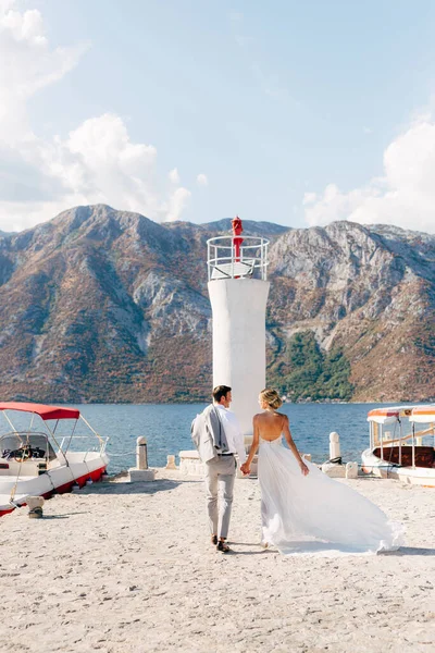 De bruid en bruidegom houden hand in hand langs de pier naar de vuurtoren in de baai van Kotor, achteraanzicht — Stockfoto