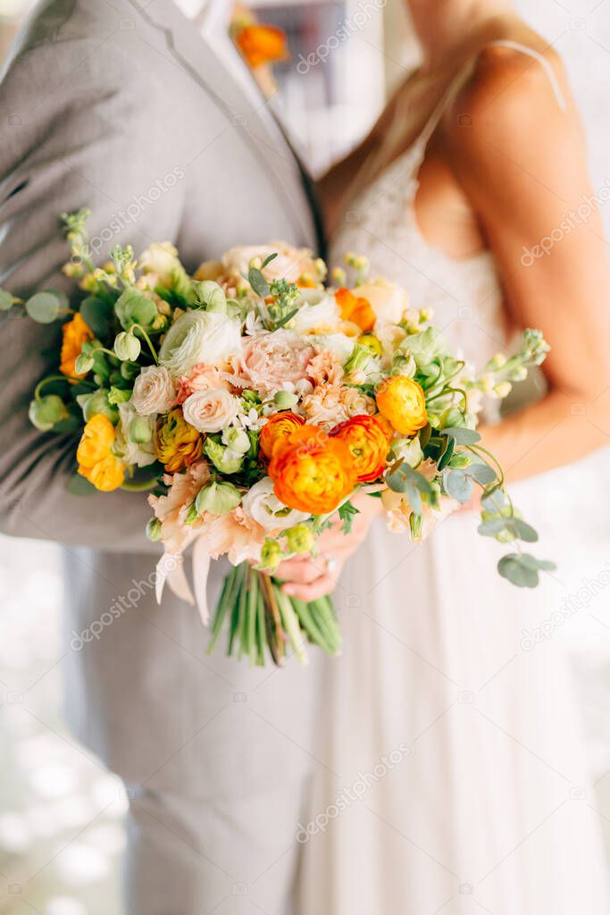 The bride and groom stand hugging and hold a wedding bouquet with orange buttercups, roses and eucalyptus branches close-up 