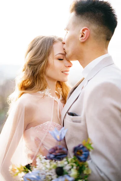 Groom kisses the forehead of smiling bride in a beautiful rose dress with a bouquet of flowers — Stock Photo, Image