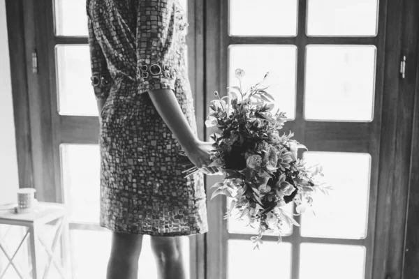 Une mariée dans une robe insolite avec des paillettes se tient à la porte du balcon et tient un bouquet de mariage, photo noir et blanc — Photo