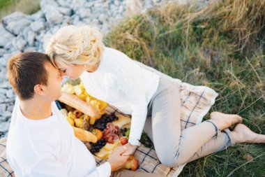 Çıplak ayaklı kadın, alnını battaniyede oturan adama yasladı. Erkek ve kadın el ele tutuşuyor. Battaniyenin üzerinde yemek var.
