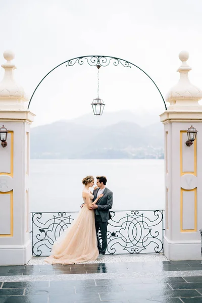 Los recién casados se encuentran en un abrazo bajo un antiguo arco apoyado en una cerca forjada contra el telón de fondo del Lago de Como —  Fotos de Stock