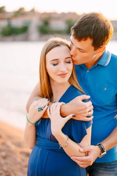 Man zachtjes zoent glimlachende zwangere vrouw in een blauwe jurk knuffelen haar schouders — Stockfoto