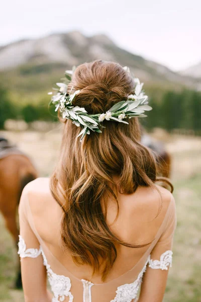 Νύφη στέκεται με την πλάτη της σε ένα όμορφο φόρεμα δαντέλα. Το κεφάλι της νύφης στολίζεται με ένα στεφάνι από πράσινα κλαδιά ελιάς. — Φωτογραφία Αρχείου