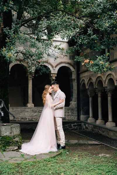 Pengantin pria memeluk pengantin wanita, berdiri di kebun hijau dengan latar belakang vila tua dengan kolom di Danau Como, Italia — Stok Foto