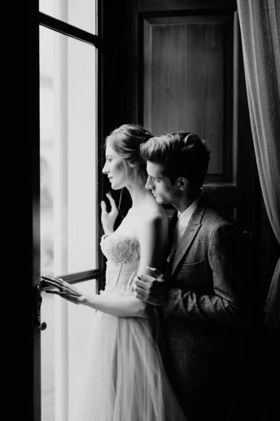 Ο γαμπρός αγκαλιάζει τη νύφη από πίσω στο μεγάλο παράθυρο μιας παλιάς βίλας. Λίμνη Κόμο. Ασπρόμαυρη φωτογραφία — Φωτογραφία Αρχείου