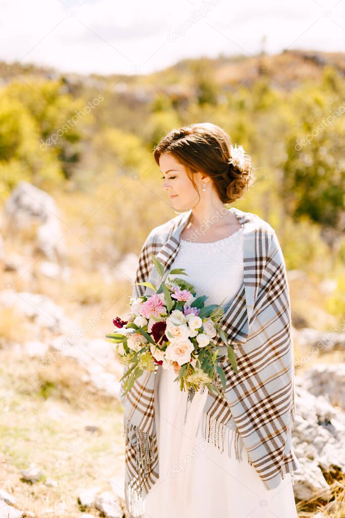 Smiling bride with a checkered shawl on her shoulders and a beautiful bouquet of flowers in her hands on a background of nature