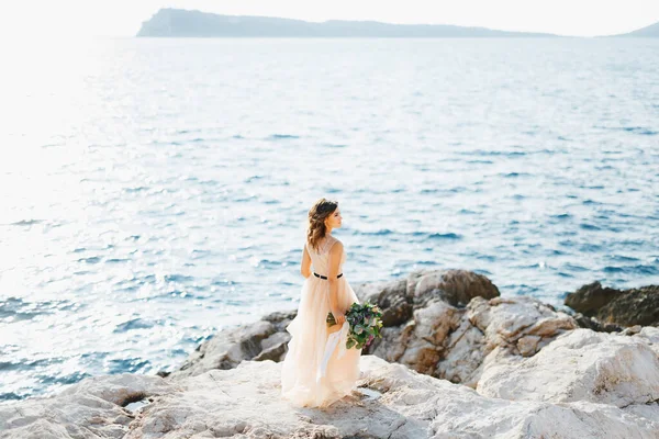 Прекрасная невеста в пастельном свадебном платье стоит на скале над морем с букетом цветов в руках — стоковое фото