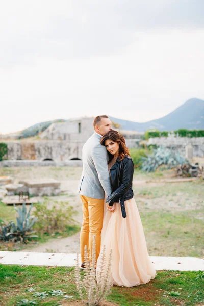 Vážné ženich a nevěsta v pastelových svatebních šatech a černá kožená bunda objímají na pozadí zříceniny hradu — Stock fotografie