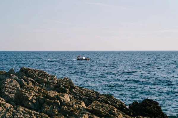 Una lancha a motor con toldo y los turistas montan las olas en el mar, cerca de la costa rocosa. — Foto de Stock