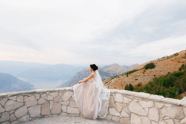 A noiva se senta no lado de pedra do deck de observação na montanha Lovchen e olha para a Baía de Kotor — Fotografia de Stock