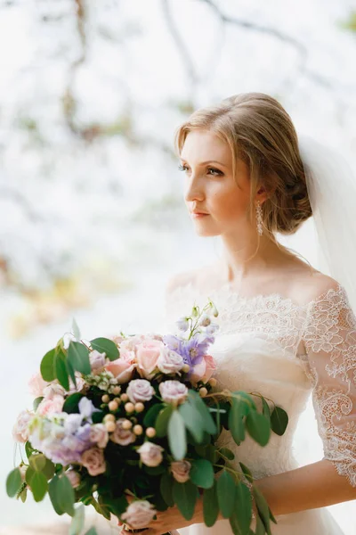 Una sposa sofisticata sta con un bouquet da sposa tra le mani sotto i rami di un albero, primo piano — Foto Stock
