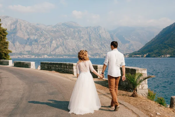 La novia y el novio caminan de la mano a lo largo de la carretera a lo largo de la costa en la bahía de Kotor cerca de Perast, vista trasera —  Fotos de Stock