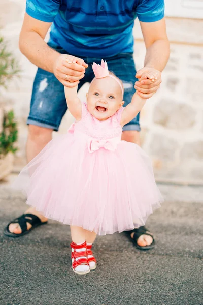 Une charmante petite fille en robe rose, baskets et avec une couronne sur la tête tenant papa par les mains et riant — Photo