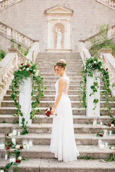 Skonsam brud med en bröllopsbukett nära pelarna dekorerade med blommor och ljus på den antika trappan i templet i Prcanj — Stockfoto