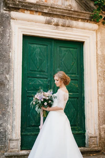 Seorang pengantin lembut berdiri dengan buket di tangannya terhadap latar belakang pintu hijau kayu dari sebuah bangunan kuno — Stok Foto
