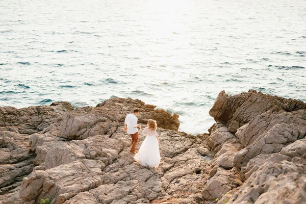 La novia y el novio se paran tomados de la mano en las rocas junto al mar y se miran entre sí —  Fotos de Stock