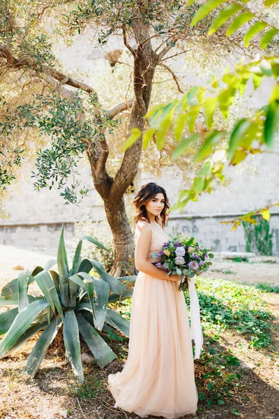 Härlig brud i en beige klänning står mot ett träd bakgrund med en vacker bukett blommor i händerna — Stockfoto