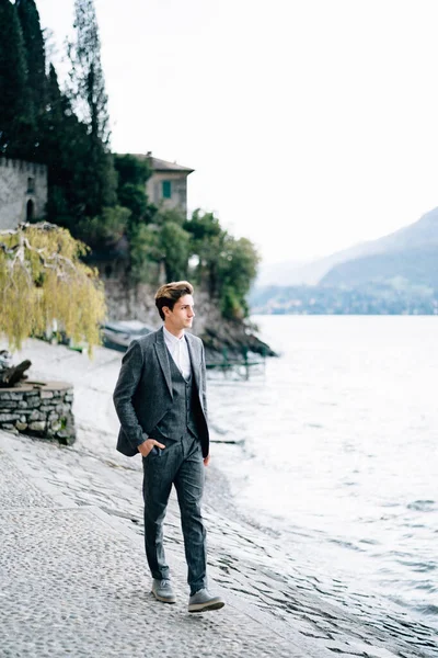 El novio camina a lo largo del terraplén de la ciudad de Varenna en el lago de Como contra el telón de fondo de edificios antiguos y montañas — Foto de Stock