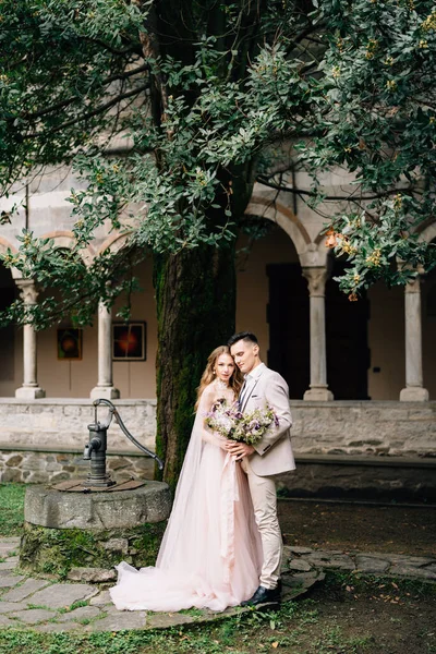 Groom embrasse mariée avec un bouquet de fleurs dans un jardin verdoyant sur fond de conduite d'eau, un arbre et un vieux bâtiment sur le lac de Côme. — Photo