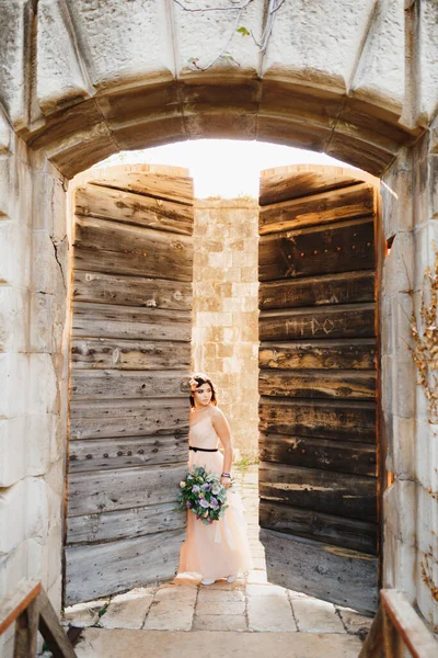 Pengantin yang cantik dengan gaun yang indah dengan karangan bunga condong ke pintu benteng yang terbuka dari kayu — Stok Foto