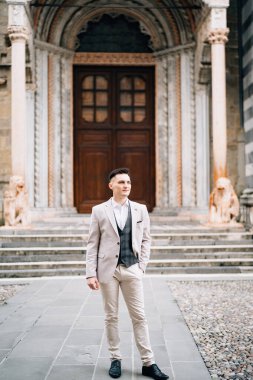Gülümseyen damat bej bir takım elbiseyle İtalya 'nın Bergamo kentindeki antik bir kapının arkasında duruyor.
