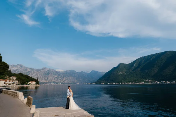 Os recém-casados estão abraçando uns aos outros no cais contra o pano de fundo do mar e montanhas verdes. Perast town, Montenegro — Fotografia de Stock