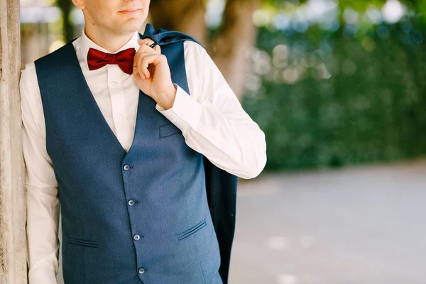 Groom holds his jacket on his shoulder, leaning against a pillar in the garden — Stock Photo, Image