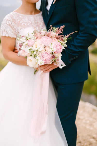 Les mariés se tiennent dans les bras et tiennent le bouquet de mariées avec de délicates roses, pivoines et astilbe, gros plan — Photo