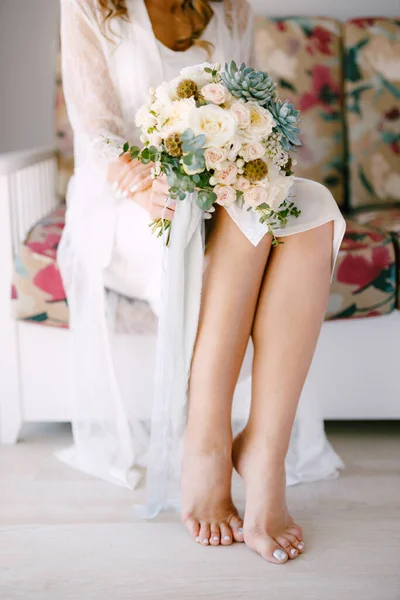 Barefoot bride in a lace robe sits on a sofa in a hotel room and holds a wedding bouquet in her hands, close-up — Stock Photo, Image