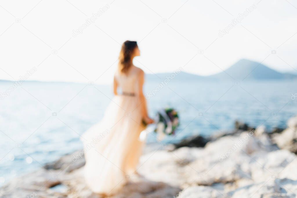 Silhouette of lovely bride in a pastel wedding dress stands on a rock above the sea with a bouquet of flowers in her hands