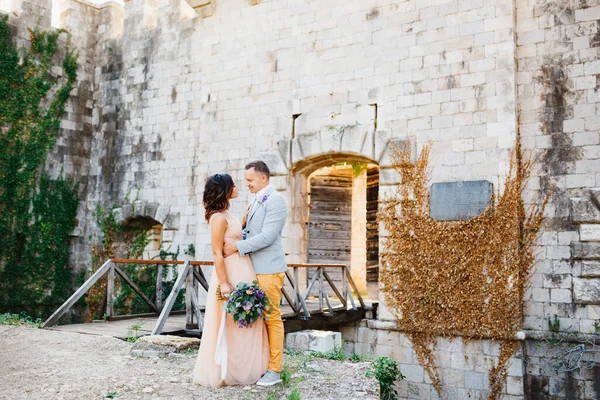 Groom étreint la mariée dans une jolie robe pastel sur fond de pont menant au château. Mariée tient un bouquet de fleurs dans ses mains — Photo