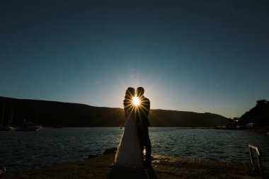 Damat ve gelin gün batımında el ele tutuşup sahilde dururlar.