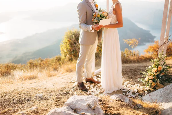 A noiva eo noivo estão abraçando no Monte Lovcen perto do arco de casamento, o noivo detém um envelope em suas mãos — Fotografia de Stock
