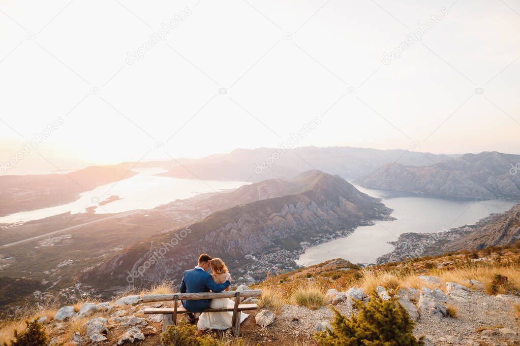 The bride and groom sitting and hugging on the wooden bench on the top of Mount Lovcen overlooking the Bay of Kotor