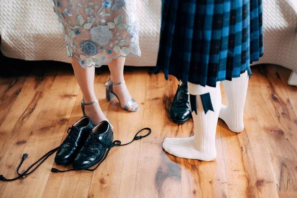 Persiapan pernikahan Skotlandia. Pria berrok pendek berdiri di samping wanita dengan sepatu hak tinggi. Ada sepatu dengan tali panjang di lantai — Stok Foto