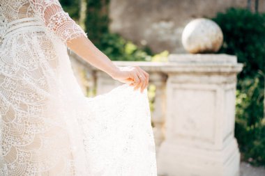 Gelin, parmaklarıyla bahçedeki sütunların arkasında beyaz dantelli bir gelinliğin zeminini tutuyor.