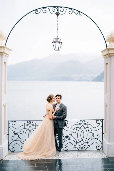 Pengantin baru memeluk dan hampir mencium di bawah lengkungan tua terhadap latar belakang Danau Como — Stok Foto