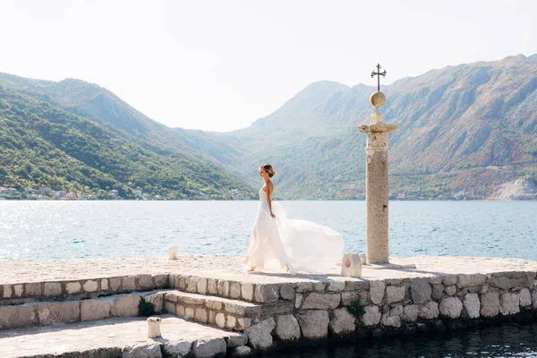 Panna młoda spaceruje po kamiennym molo obok starej kolumny w Zatoce Kotor, jej spódnica trzepocze na wietrze — Zdjęcie stockowe