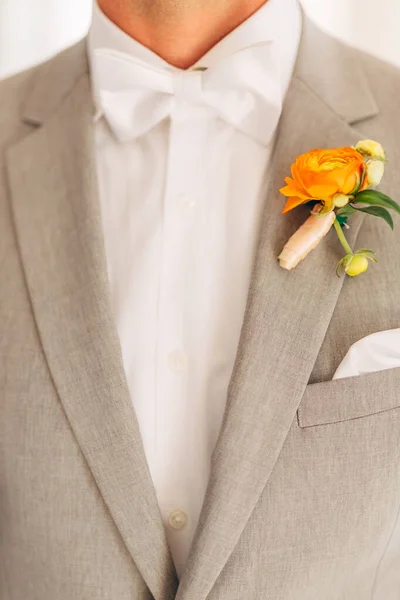 Le marié en costume gris avec un nœud papillon blanc et une boutonnière aux fleurs orange dans une chambre d'hôtel, gros plan — Photo