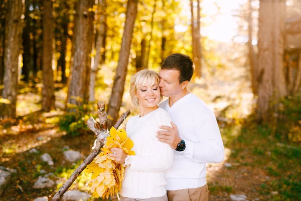 Mann küsst lächelnde Frau vor dem Hintergrund eines Herbstwaldes. Frau hält einen Strauß gelber Blätter in der Hand — Stockfoto