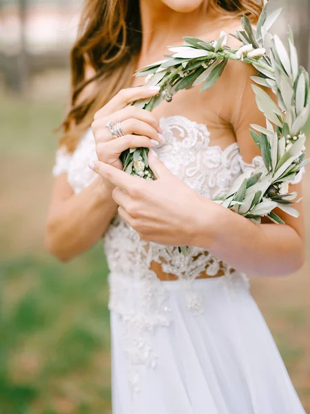 Bela noiva em um vestido branco bordado fica de lado e mantém uma coroa de ramos de oliveira verde em suas mãos — Fotografia de Stock