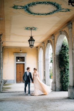 Gelin ve damat eski terasta yeşil sarmaşıklarla sarmalanmış sütunlarla yürürler. Como Gölü, İtalya. Ön görünüm