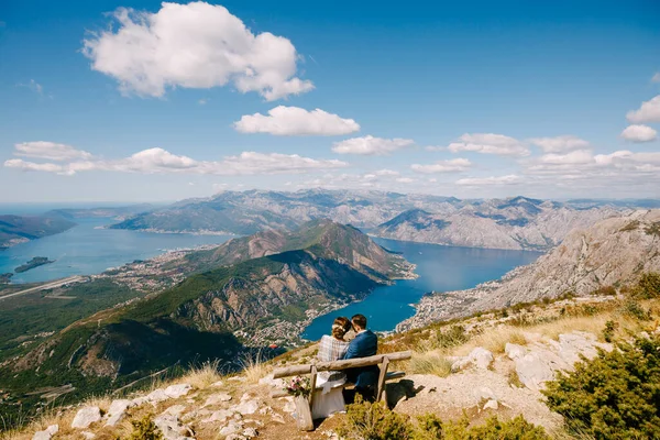 Noiva e noivo estão sentados em um banco e olhando para a baía de Kotor. Excelente vista das montanhas de Montenegro — Fotografia de Stock