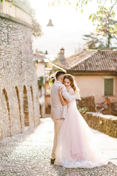 Pengantin pria memeluk pengantin wanita dengan gaun yang indah dengan karangan bunga di latar belakang bangunan kuno di Bergamo, Italia — Stok Foto