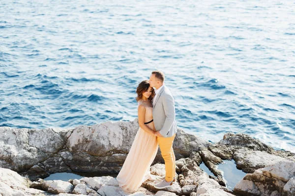 El novio sostiene las manos de las novias mientras está de pie sobre las rocas sobre el mar azul. Vista desde arriba —  Fotos de Stock