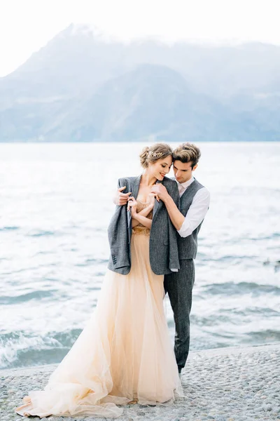Жених бросил куртку через плечи невесты, обнимающей ее на набережной озера Комо у воды — стоковое фото